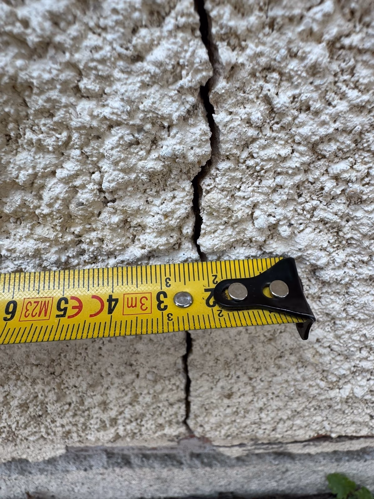 Mètre ruban tenu en travers d'une fissure verticale ouverte dans un enduit de façade granuleux, au-dessus du soubassement