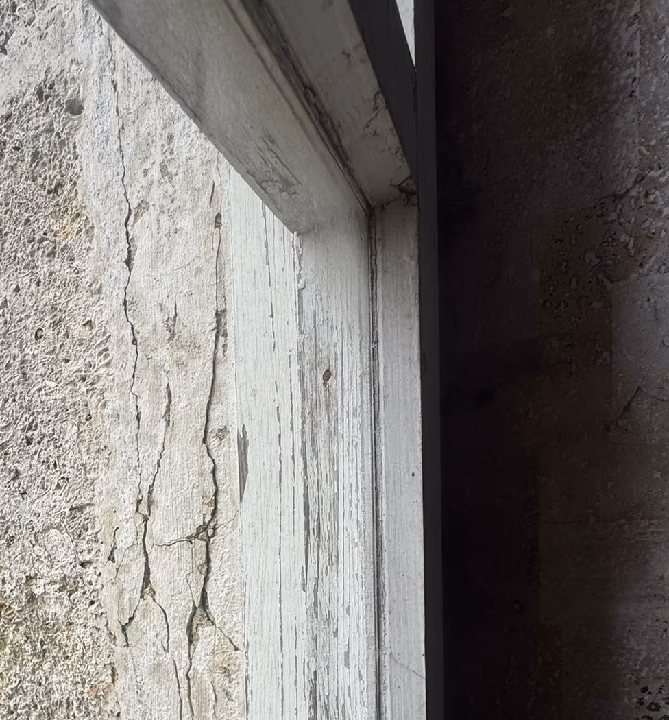Tableau extérieur d'une fenêtre ancienne : enduit peint fendu en réseau et soulevé par plaques contre un dormant en bois dont la peinture s'écaille