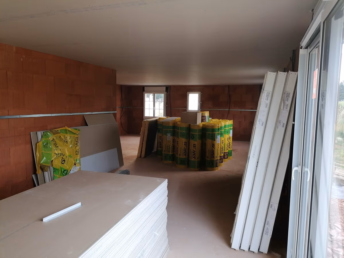 Matériaux stockés dans une maison en chantier : pile de plaques de plâtre, rouleaux d'isolant et menuiseries emballées appuyées contre le mur