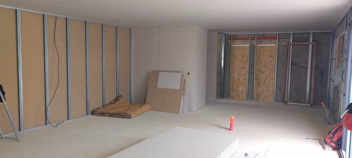 Vue d'ensemble d'un plateau de maison neuve en cours de cloisonnement : ossatures métalliques montées, doublages en partie posés, plaques de plâtre stockées au sol