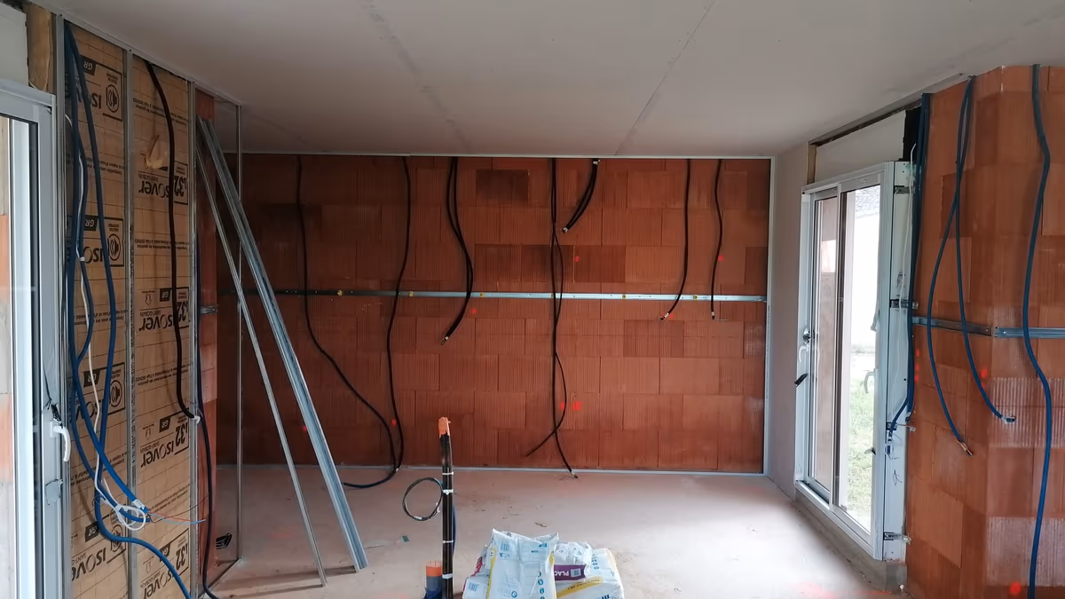 Pièce de maison neuve laissée en l'état : murs en briques nus, gaines électriques pendantes en attente, rail de doublage posé et sacs de produit au sol