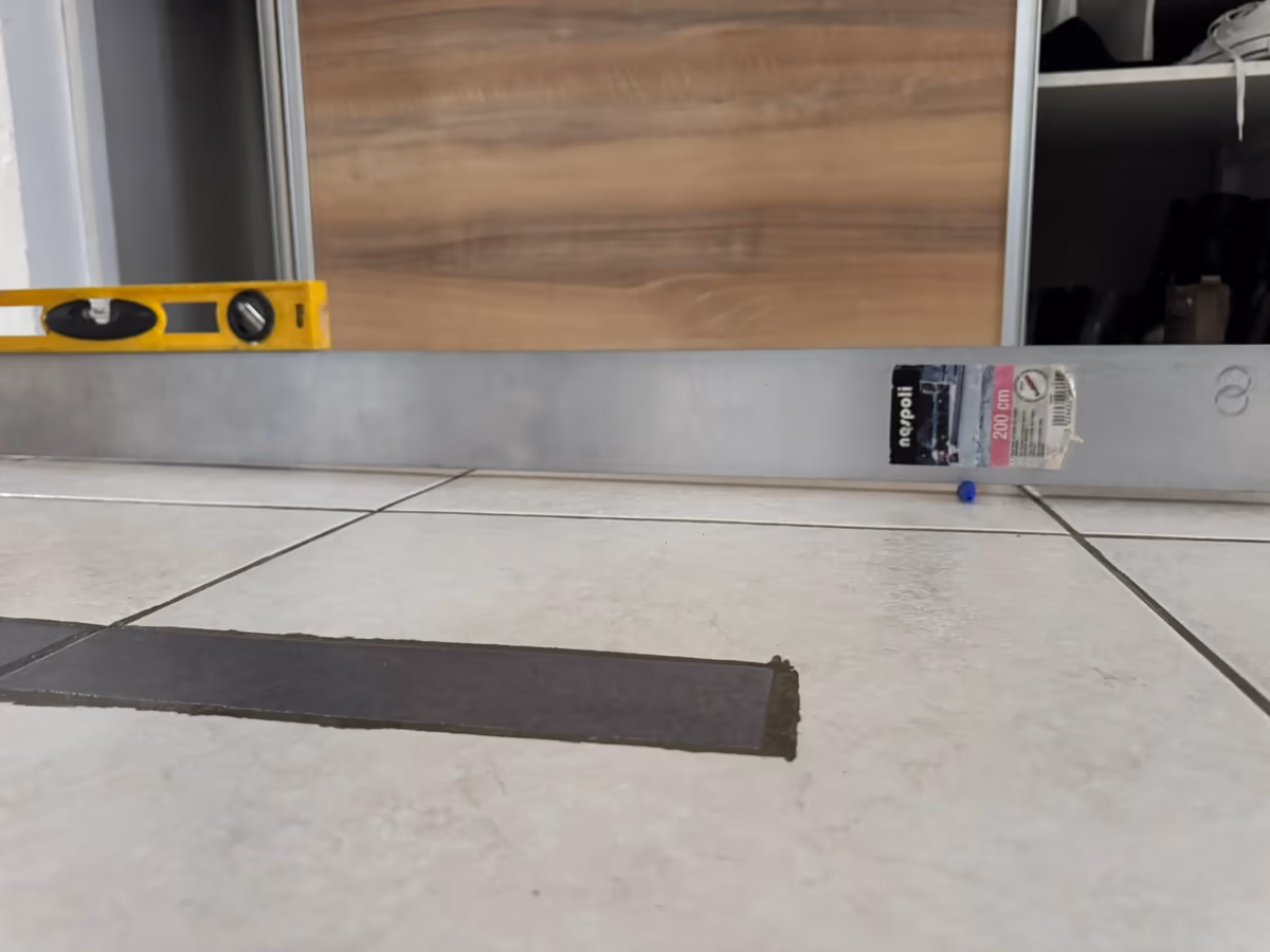 Règle de maçon en aluminium posée à plat sur un carrelage clair, niveau à bulle appuyé dessus, l'écart sous la règle observé au ras du sol