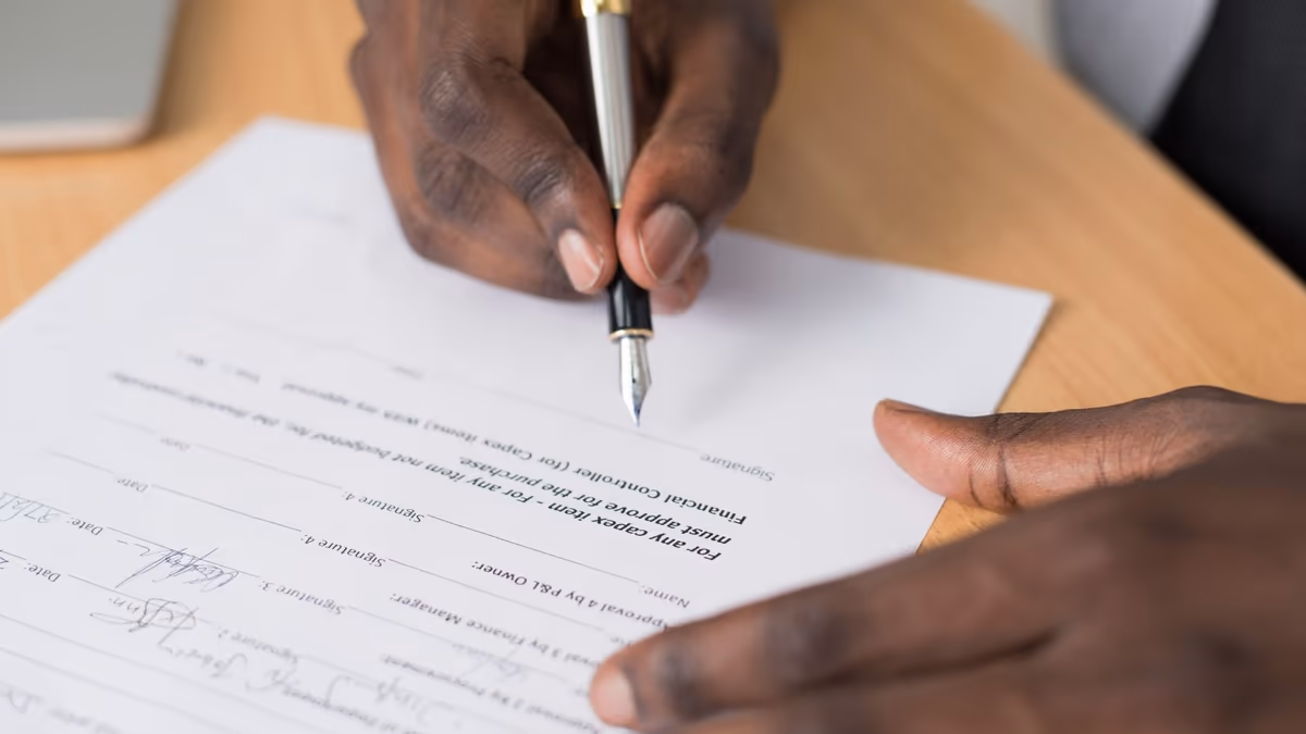 Main signant un acte de vente immobilière au stylo plume, pages de clauses et lignes de signature visibles sur la table