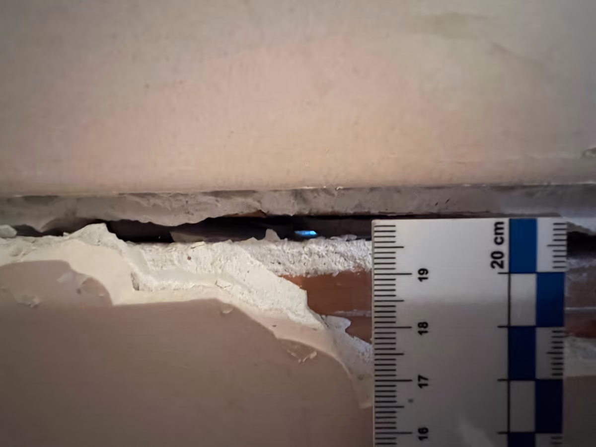 Réglette graduée en centimètres posée contre une fissure ouverte dans un revêtement mural intérieur, la brique visible au fond de l'éclat
