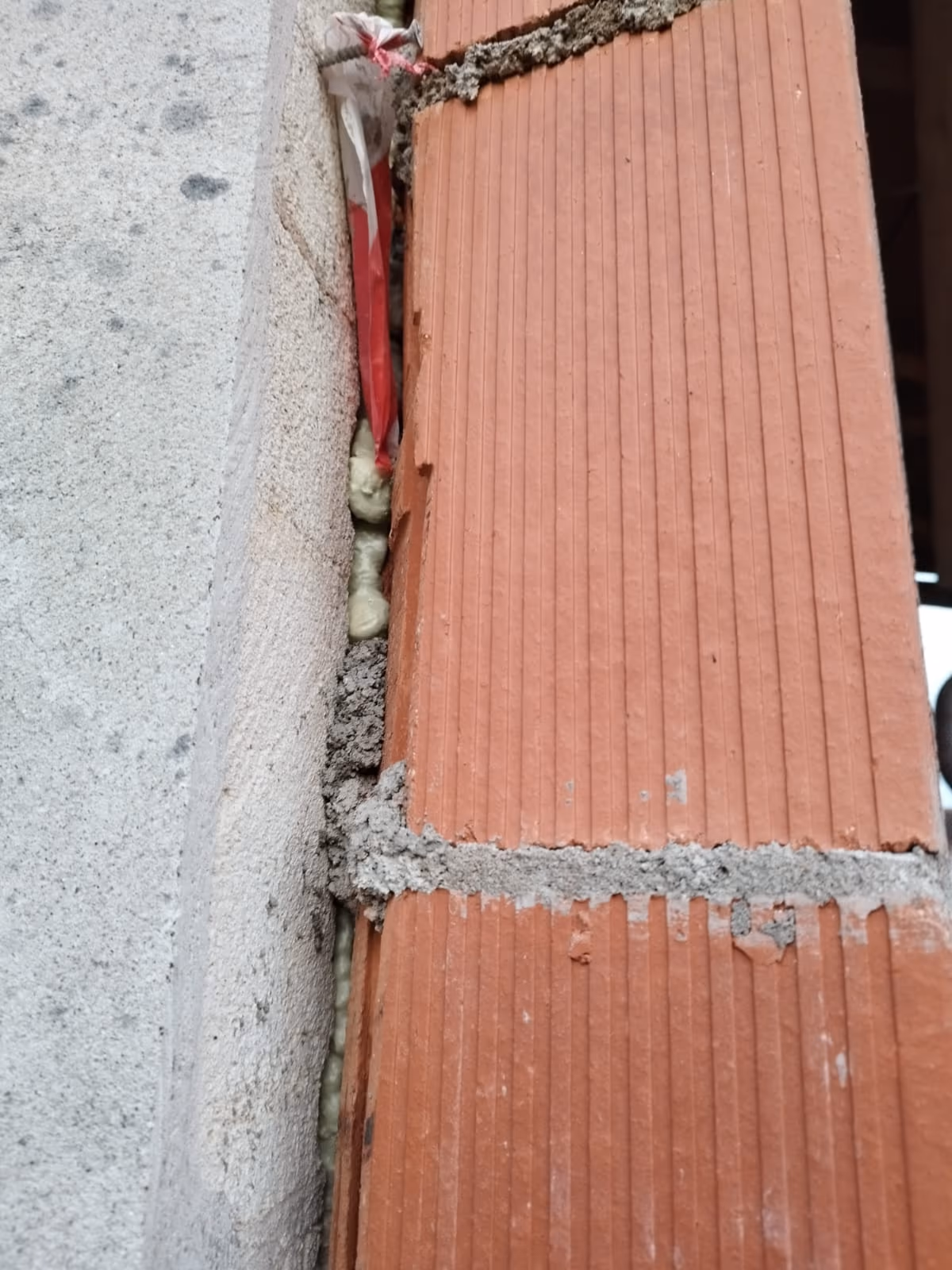 Jonction verticale ouverte entre le mur en briques d'une extension et la maçonnerie enduite existante, garnie de mousse et de mortier par endroits