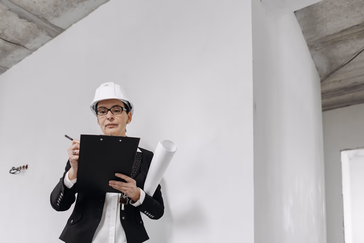Expert en casque de chantier rédigeant son constat sur un porte-bloc, plan roulé sous le bras, pendant la visite d'un logement