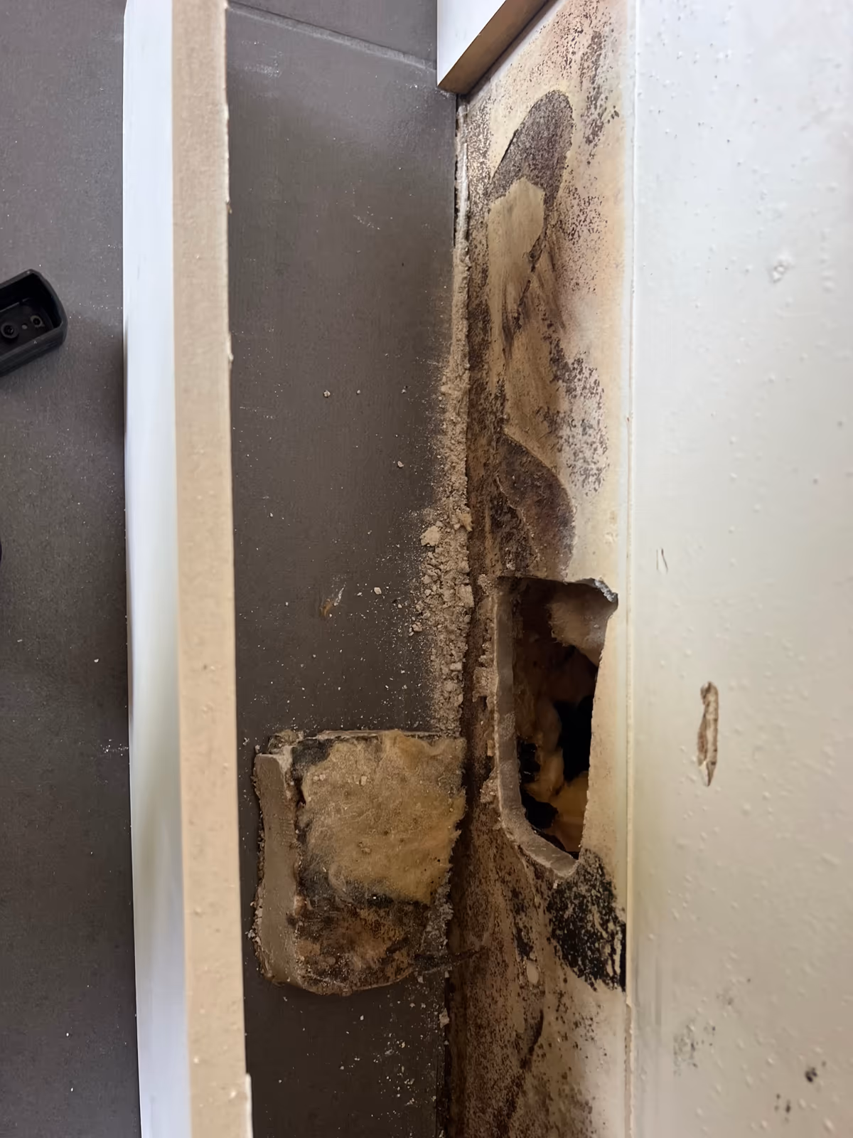Angle de cloison entaillé en pied, morceau de doublage retiré avec son isolant noirci, parement couvert de moisissures au-dessus de l'ouverture