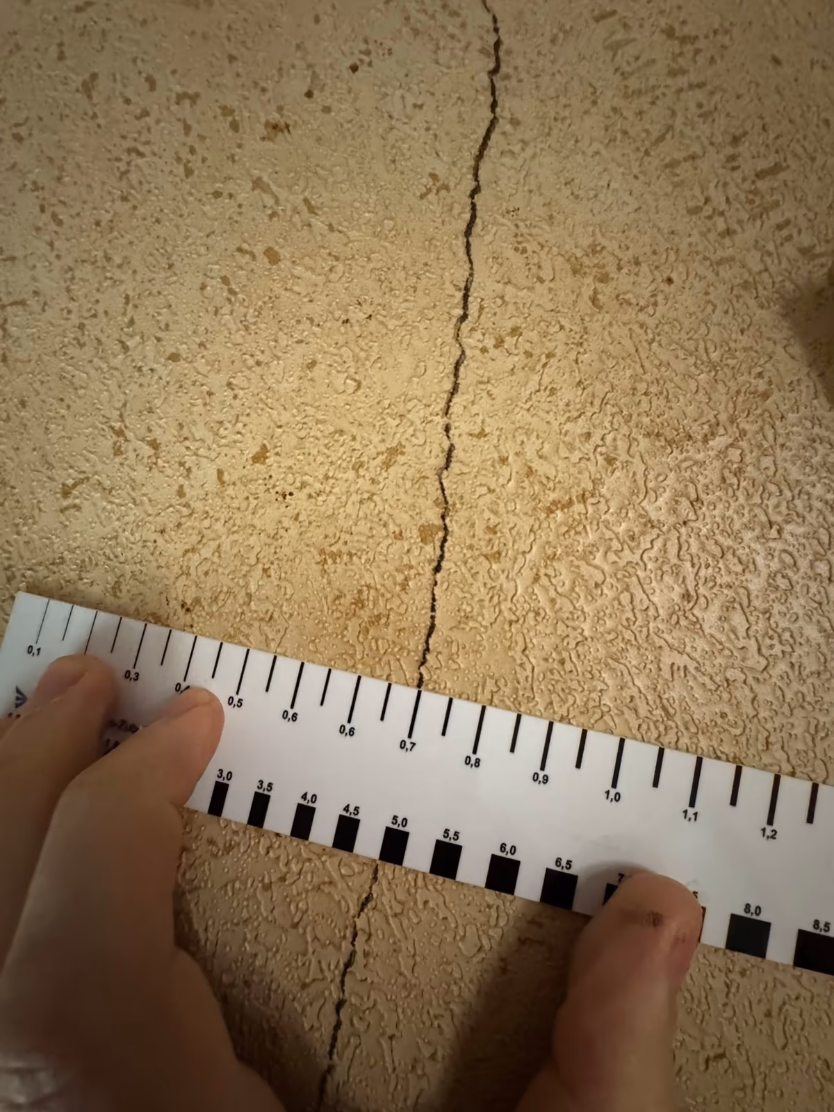 Réglette graduée tenue à deux mains en travers d'une fissure verticale sur un revêtement mural crépi, pendant un relevé sur place