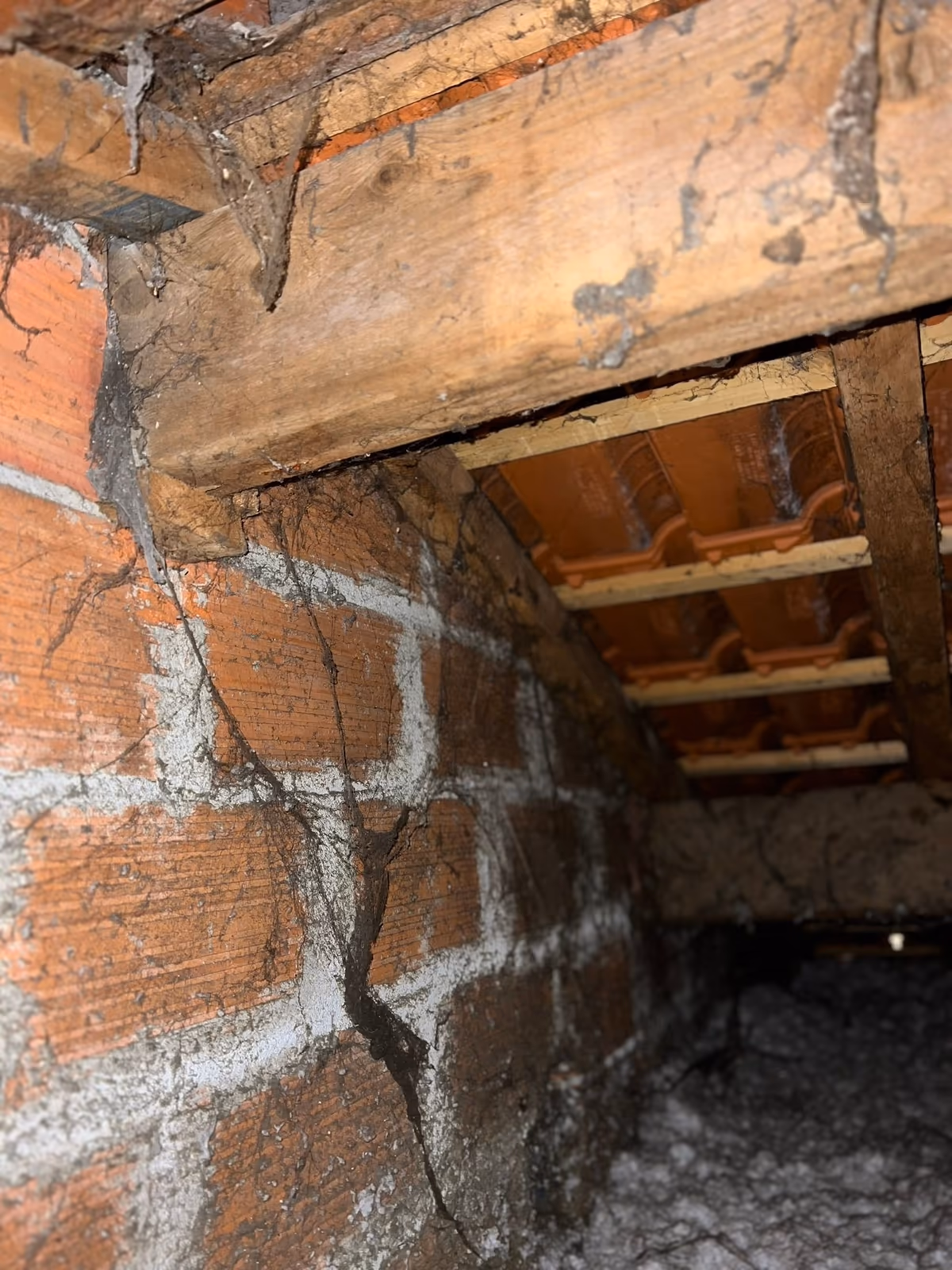 Mur en briques d'un comble fissuré sur toute sa hauteur sous une panne en bois, toiles d'araignée et isolant au sol