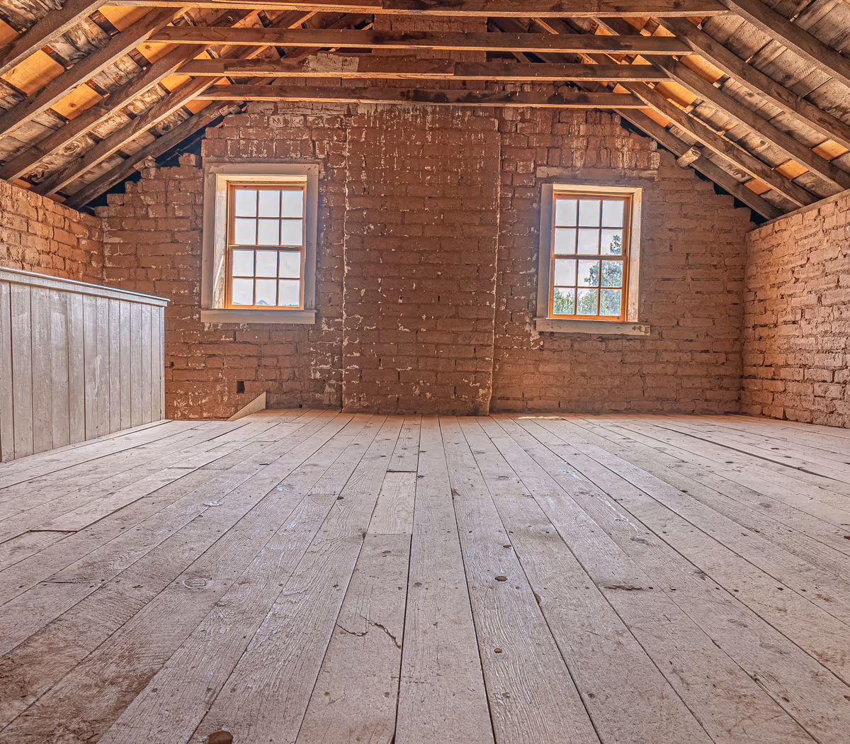 Combles d'une maison ancienne : plancher en larges planches de bois brut, pignon en brique et chevrons de charpente apparents sous la couverture