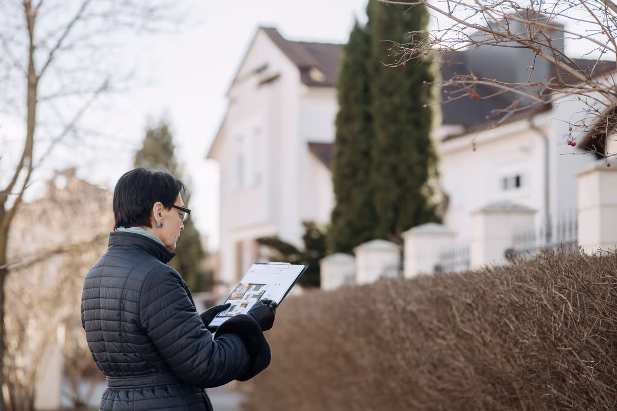 Personne debout dans la rue devant une maison, dossier ouvert à la main, observant la façade et la toiture avant une visite