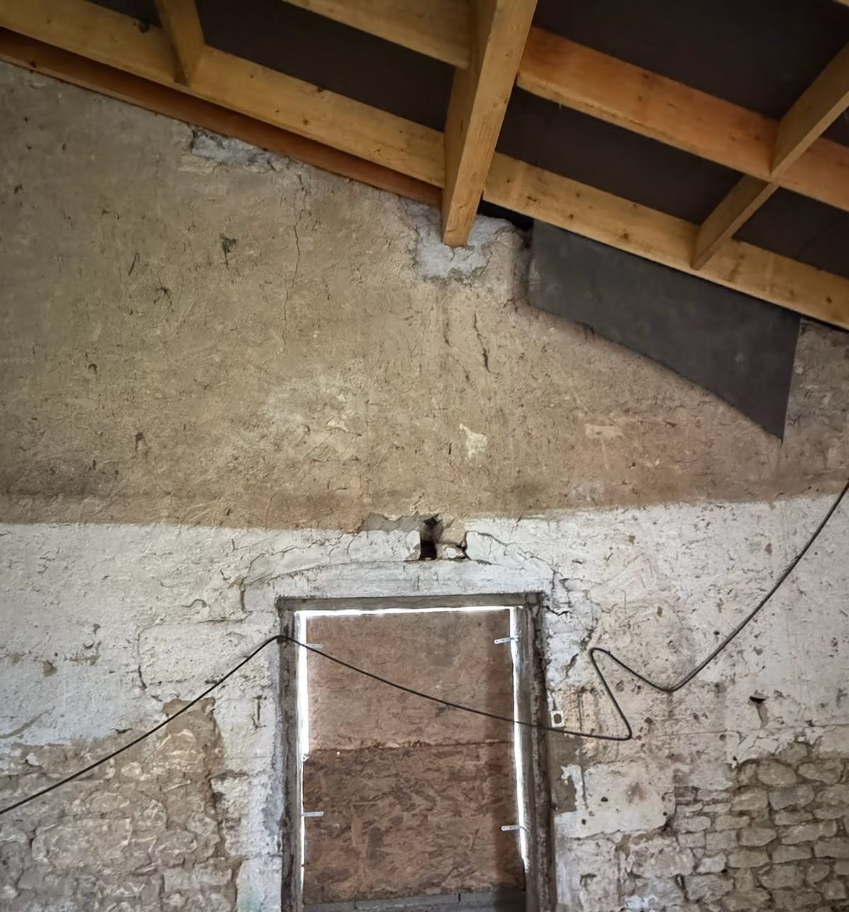Mur intérieur ancien en moellons sous une charpente neuve, enduit lacunaire, fissures en partie haute et ancienne ouverture obturée par un panneau de bois