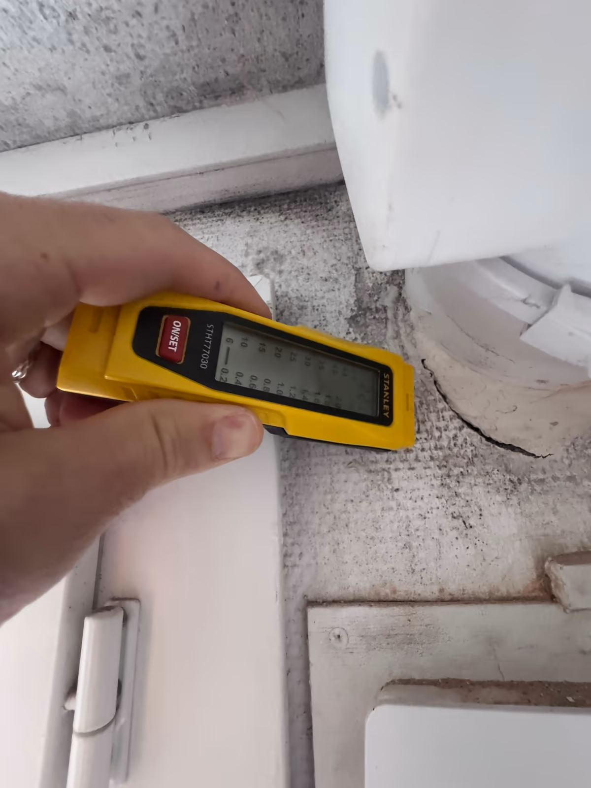 Humidimètre appliqué sur un support piqué de moisissures, dans un angle contre une menuiserie dont on voit la paumelle