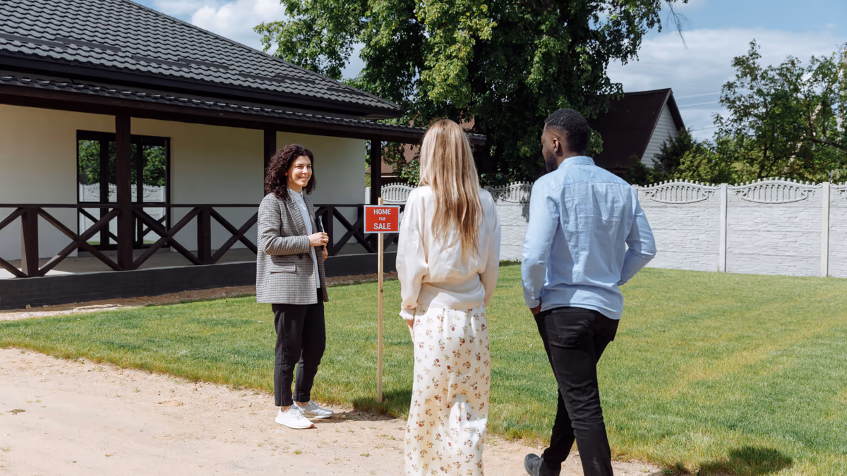 Couple d'acquéreurs arrivant devant une maison individuelle mise en vente, panneau « à vendre » planté dans le jardin