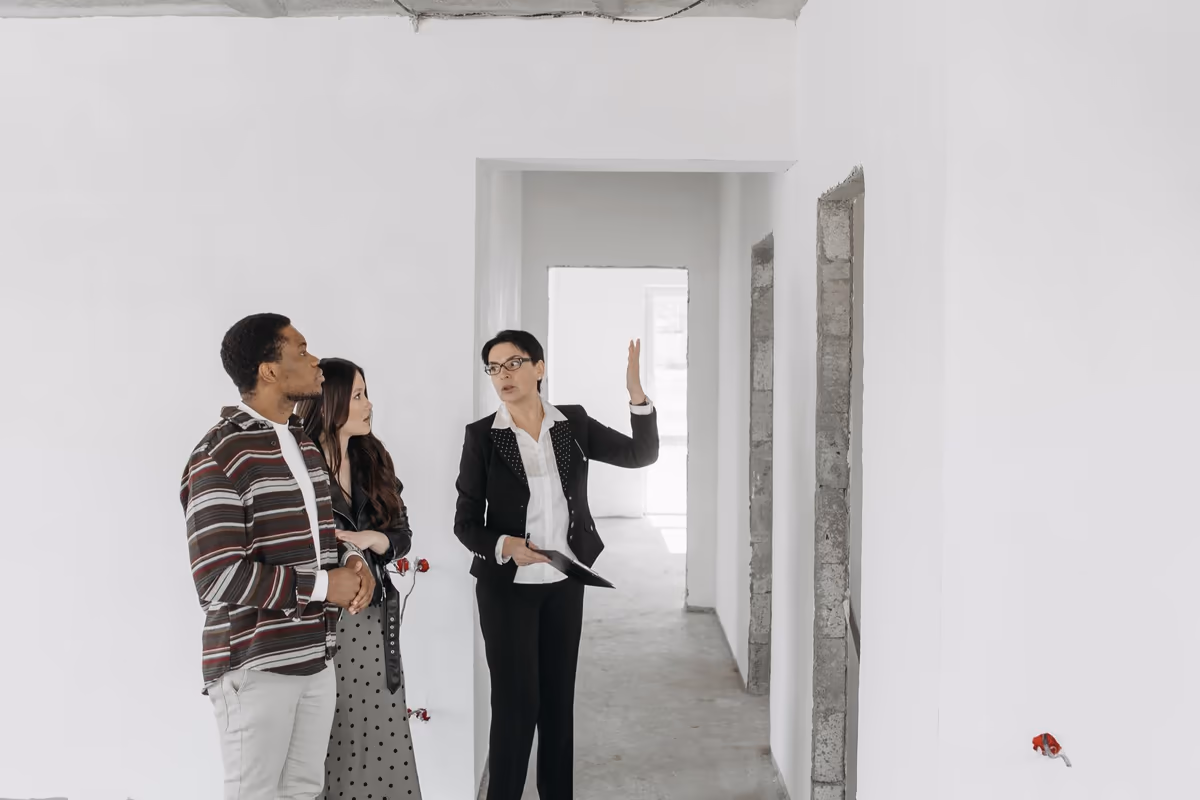 Visite commentée d'un logement vide : une professionnelle désigne un point du plafond, presse-papiers en main, tandis que les deux acquéreurs suivent son geste