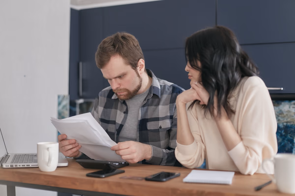 Deux acquéreurs relisant ensemble un document imprimé à la table de la cuisine, ordinateur portable ouvert à côté
