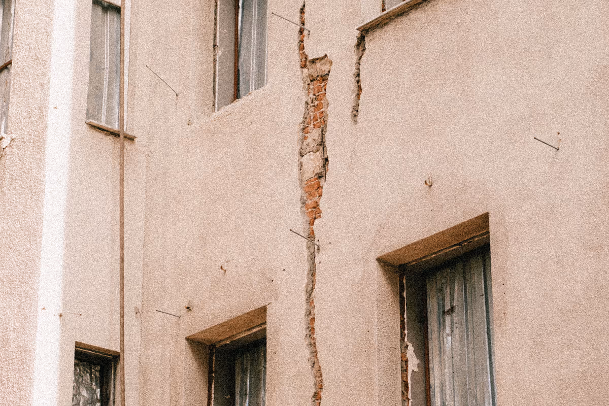 Fissure verticale traversante dans l'enduit d'une façade, filant entre deux baies et laissant apparaître la maçonnerie de brique sous-jacente