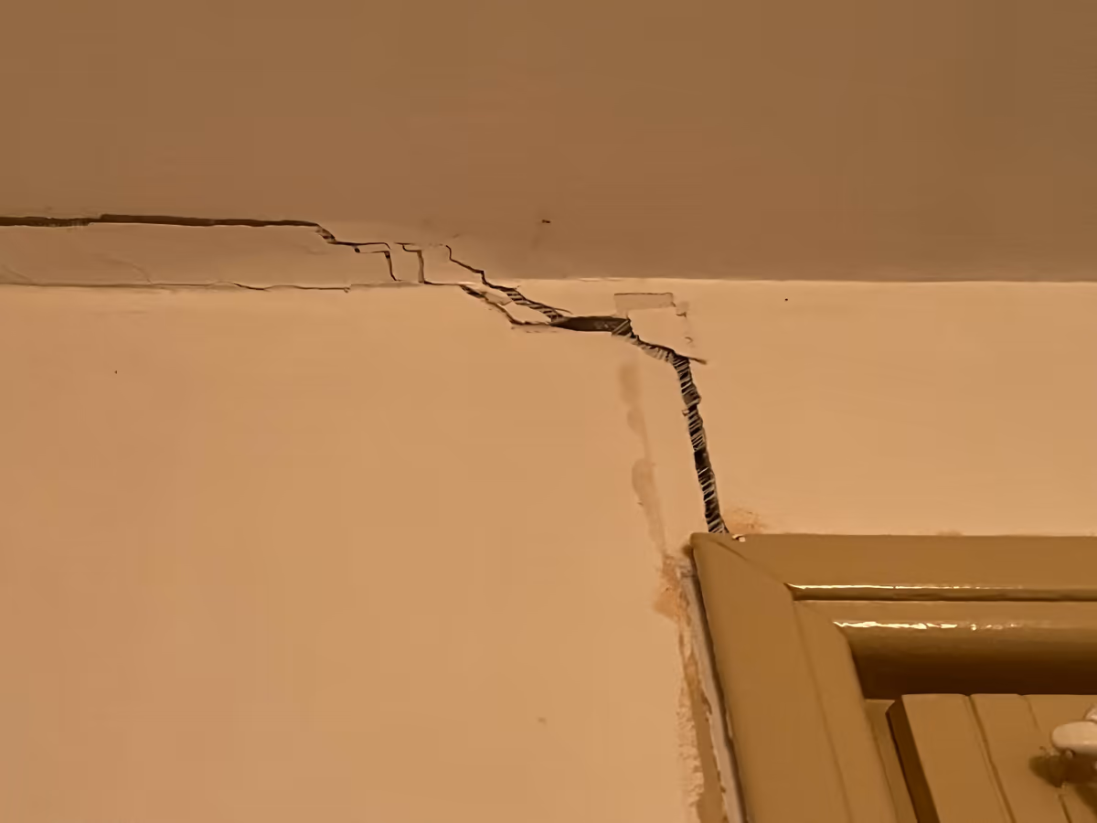Fissure largement ouverte partant de l'angle haut d'une huisserie de porte puis filant le long de la jonction entre mur et plafond