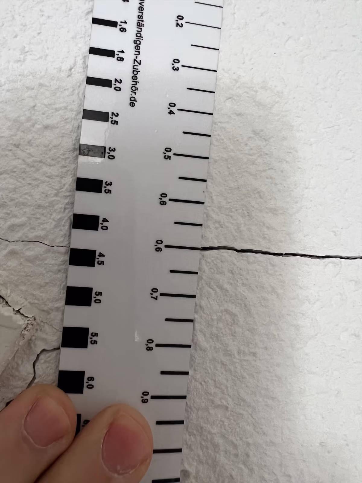 Réglette de fissuromètre tenue en travers d'une fissure horizontale très fine dans un enduit, les traits calibrés de la réglette posés juste à côté du tracé