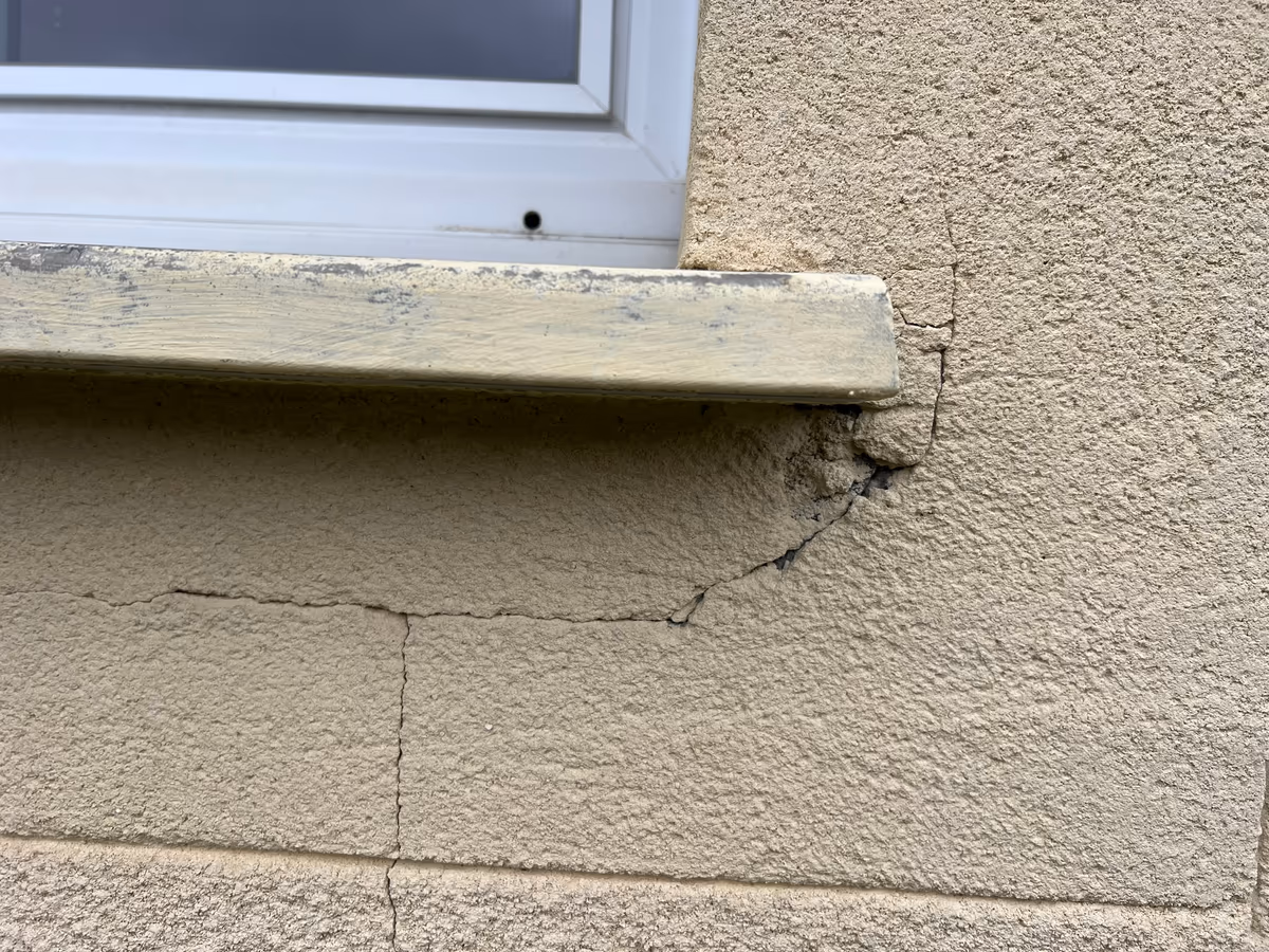 Fissures partant de l'about d'un appui de fenêtre dans un enduit de façade, l'une en arc sous l'appui, les autres suivant les joints des blocs en dessous