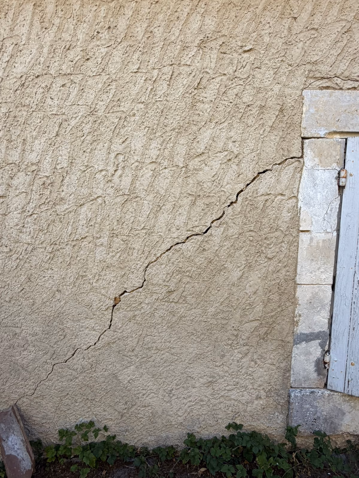 Fissure oblique qui part de l'angle d'un encadrement de fenêtre en pierre et descend en diagonale dans l'enduit de la façade jusqu'au pied du mur