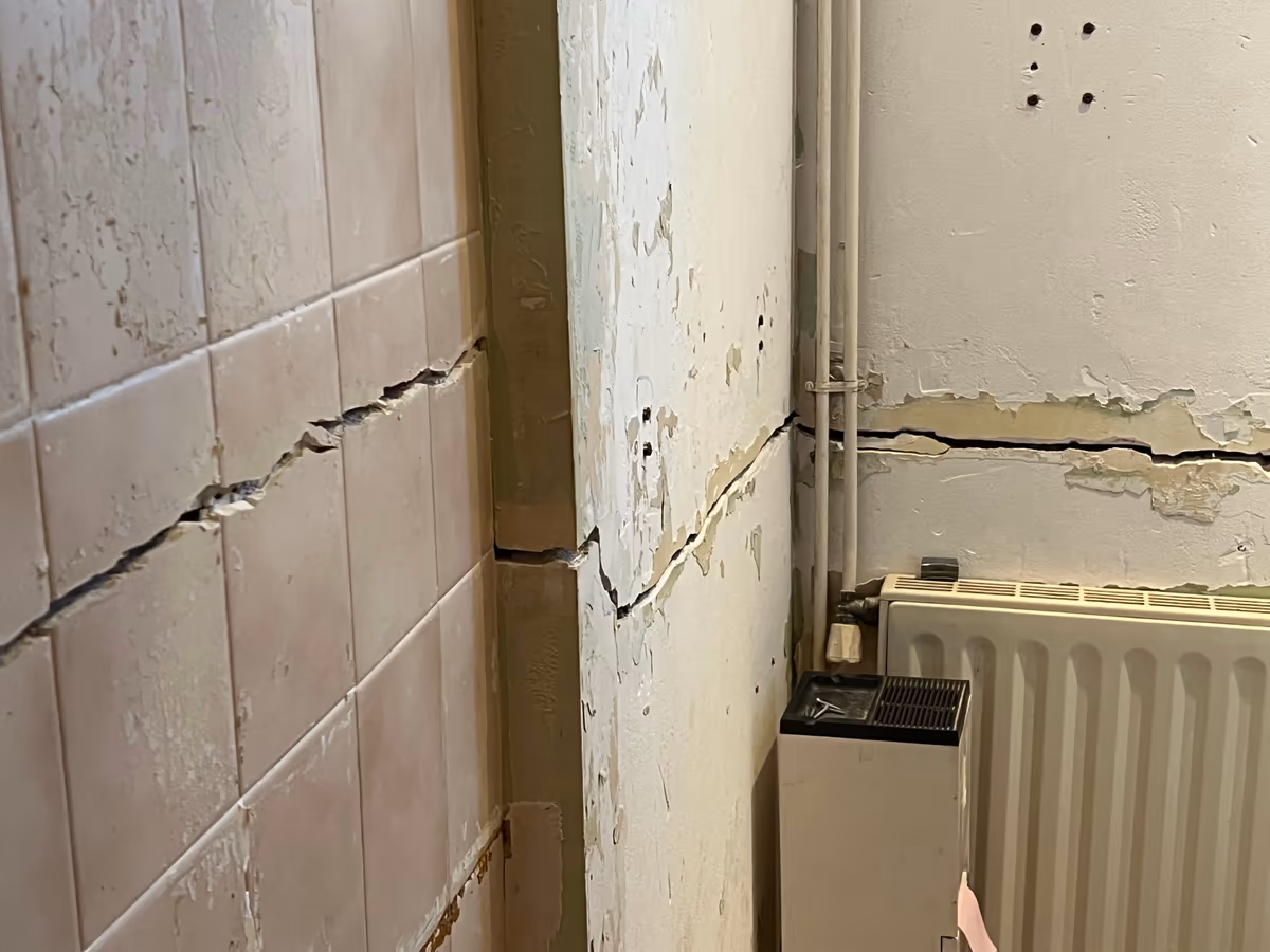 Fissure ouverte et quasi horizontale qui traverse une faïence murale, se poursuit dans l’angle d’un poteau enduit puis court sur le mur voisin au-dessus d’un radiateur