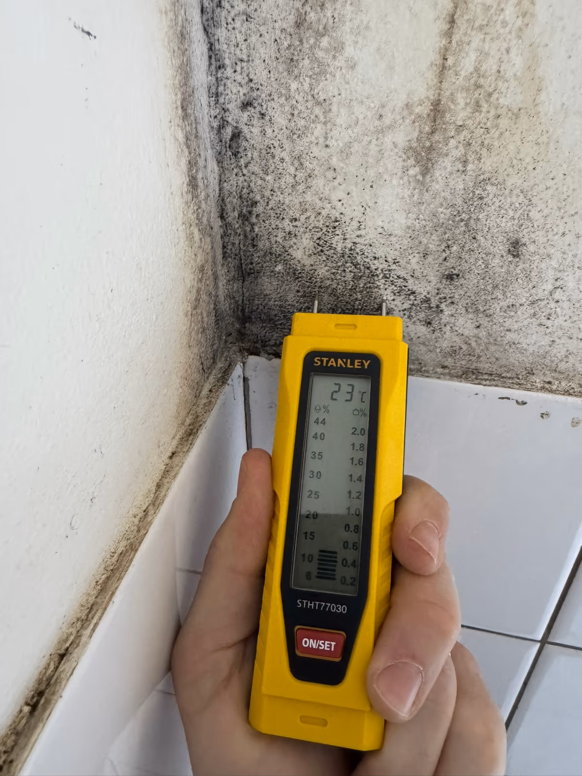 Humidimètre tenu contre un angle de mur noirci de moisissures, au-dessus d'un rang de faïence blanche