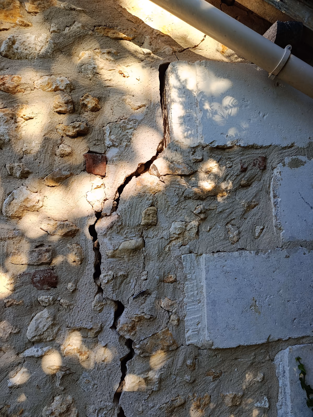 Fissure ouverte dans un mur de moellons hourdé et enduit, qui longe le bord d'une pierre de taille sous la gouttière puis descend en zigzag dans le mortier entre les moellons