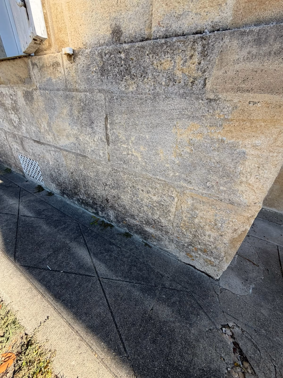 Soubassement en pierre de taille d'un angle de maison, noirci et taché sur ses assises basses au contact d'un trottoir en dalles, avec une grille d'aération au ras du sol