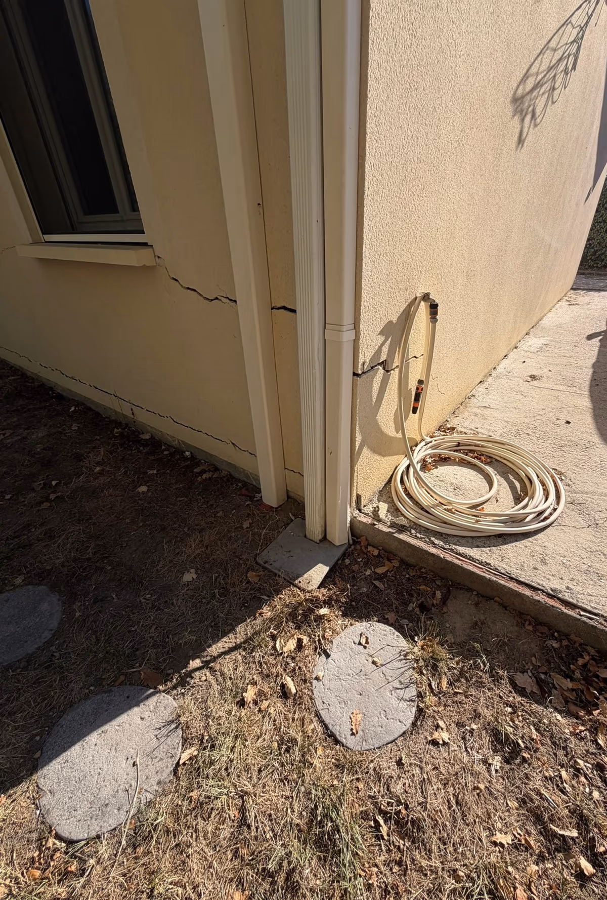 Angle de maison enduite avec deux descentes d'eaux pluviales posées sur un carreau au sol, fissures horizontales dans l'enduit sous l'appui de fenêtre et en pied de façade, terrain sec et herbe grillée au pied du mur