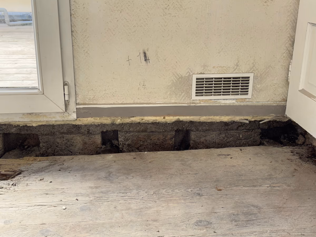 Pied de mur intérieur avec le plancher bois déposé sur une bande : soubassement maçonné à nu, réservations vides des anciens appuis de bois, grille de ventilation au-dessus