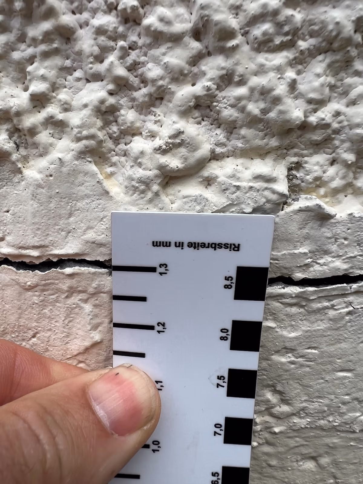 Carte fissuromètre tenue à la main contre une fissure horizontale d'enduit, ses traits d'épaisseur graduée placés au droit de l'ouverture