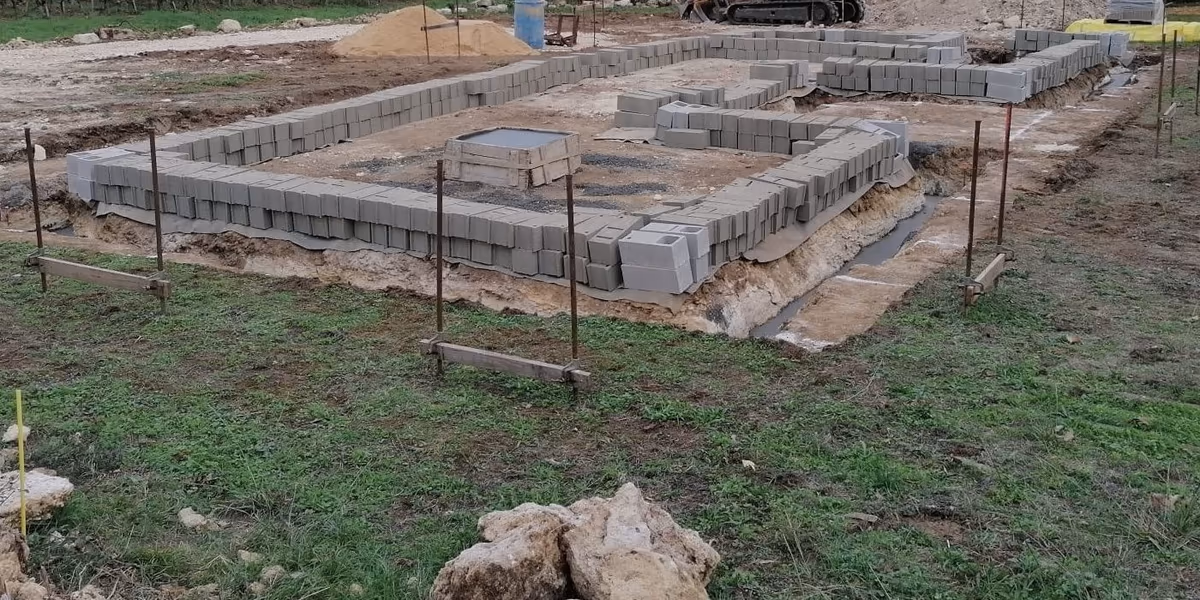 Fondations d'une maison neuve en cours : premiers rangs de blocs béton posés sur les semelles, fouilles encore ouvertes et chaises d'implantation