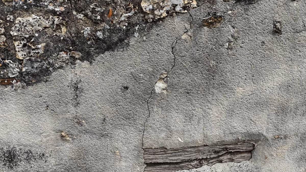 Fissure fine et sinueuse dans l'enduit d'un mur ancien, maçonnerie de moellons apparente au-dessus et linteau en bois en partie basse