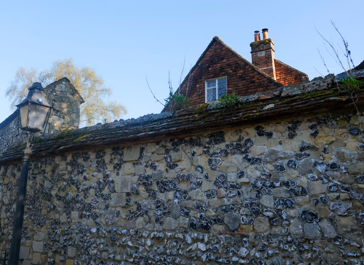 Ancien mur de clôture en silex et pierre couronné d'un chaperon de tuiles, pignon en brique d'une maison voisine avec fenêtre et cheminée au-dessus du mur