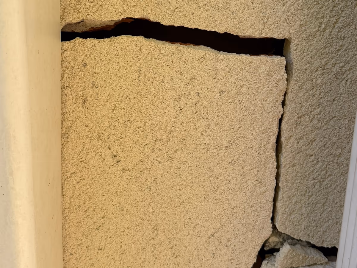 Fissure largement ouverte dans l'enduit d'une façade, au tracé décroché horizontal puis vertical, avec des morceaux d'enduit détachés à sa base