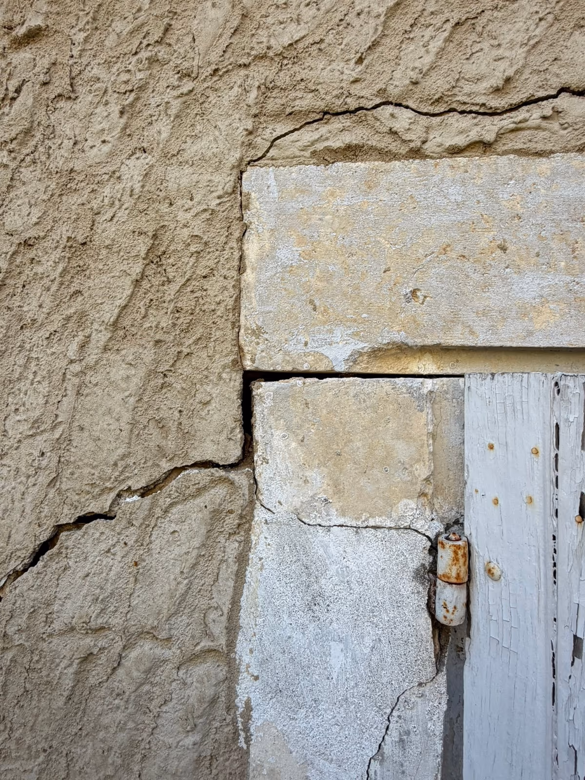 Angle d'une porte à encadrement de pierre dans un mur enduit : une fissure part du coin du linteau et descend en biais dans l'enduit, une autre court au-dessus du linteau et une troisième traverse le jambage