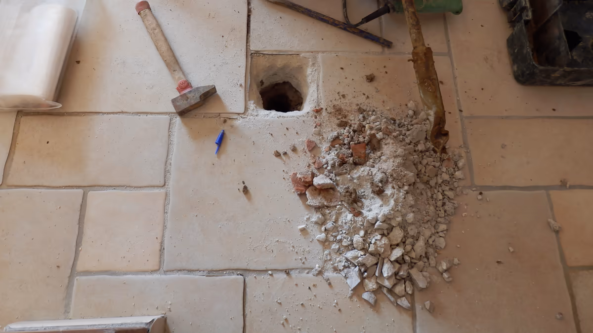 Sondage ouvert à travers un carrelage et sa dalle, gravats de la dalle à côté du trou, massette, burin et tarière posés au sol