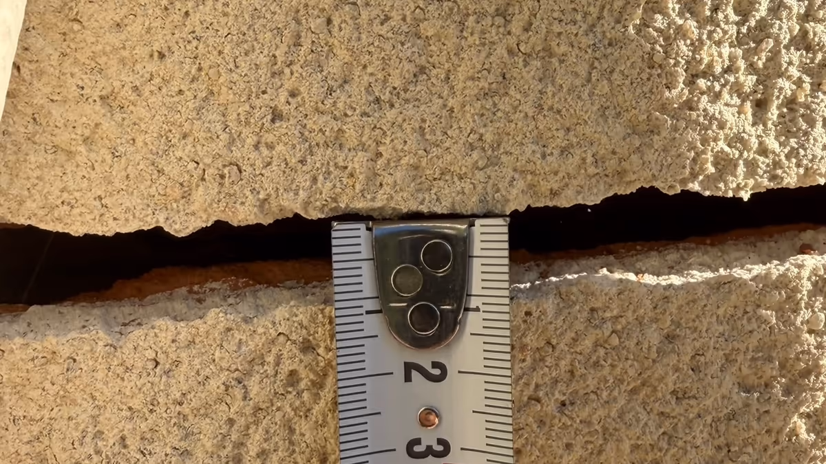 Mètre ruban posé en travers d'une fissure horizontale largement ouverte dans un enduit de façade, en plein soleil