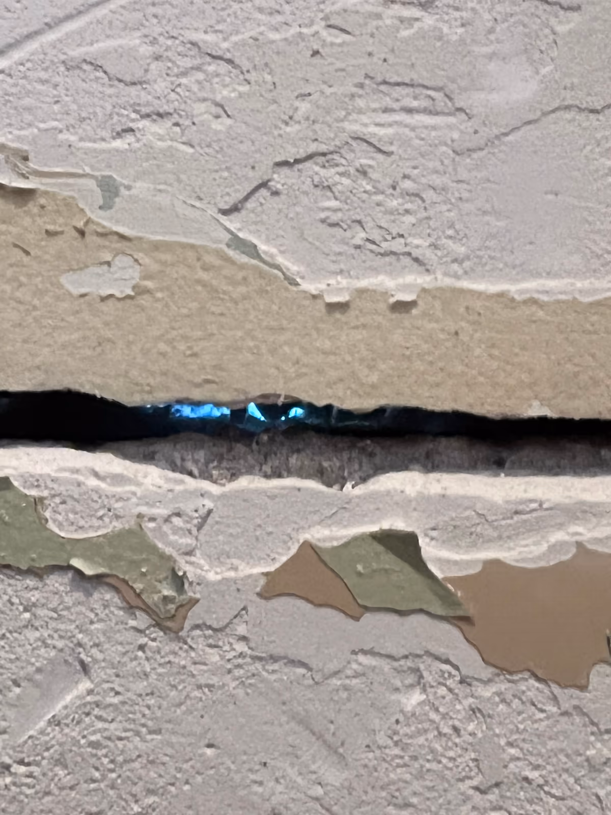 Fissure horizontale béante dans un mur intérieur, bords écaillés laissant voir plusieurs couches de peinture et d'enduit superposées