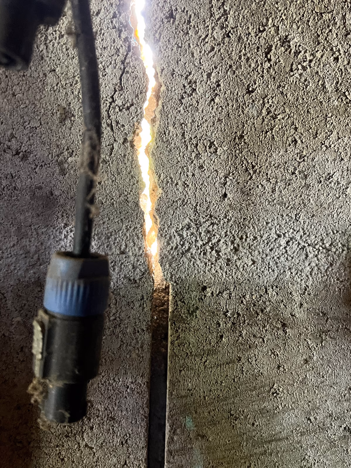 Fissure traversante dans un mur en blocs béton, le jour passant à travers l'ouverture, câble électrique pendant le long du mur