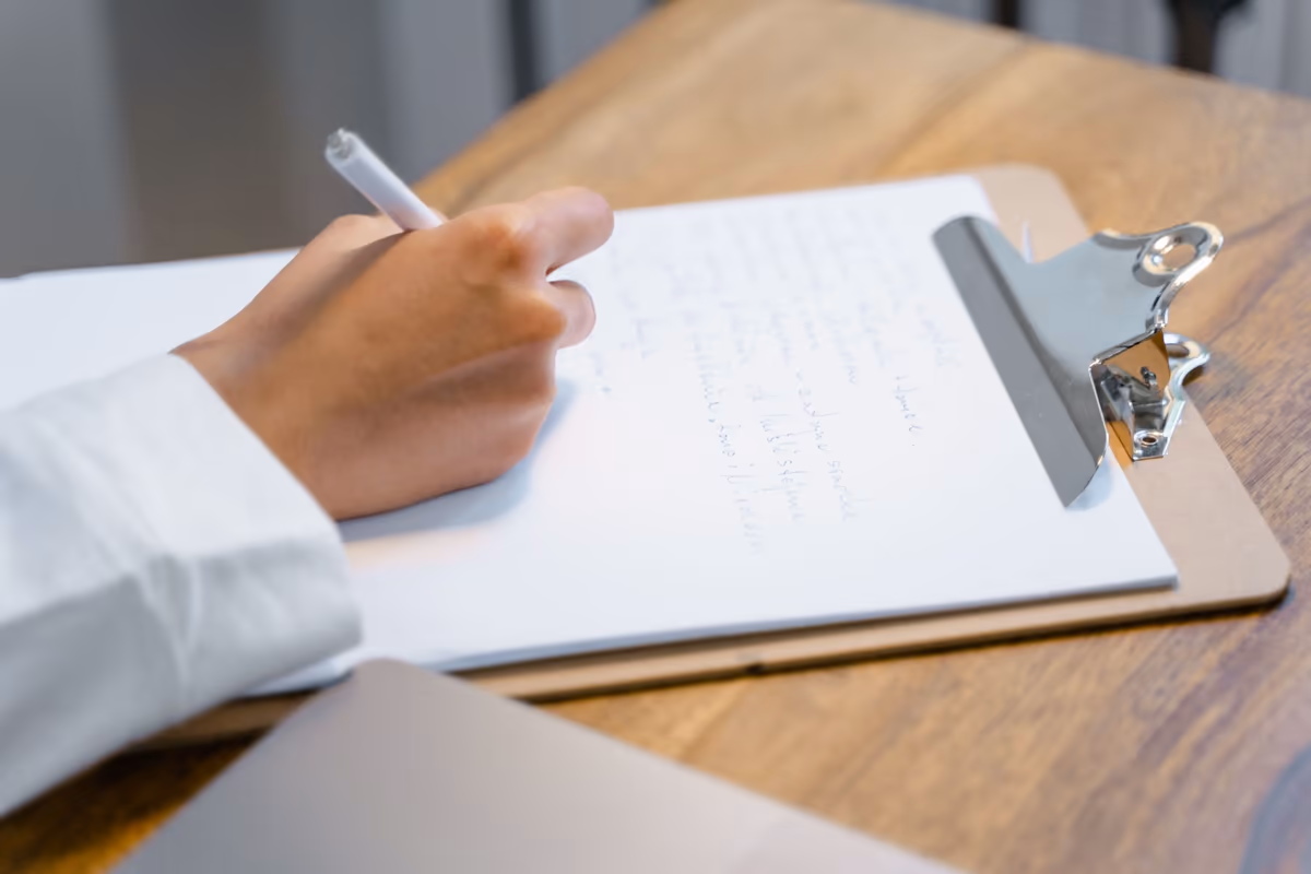 Main écrivant des observations manuscrites sur le porte-bloc d'un constat, feuillets et ordinateur posés sur la table de travail