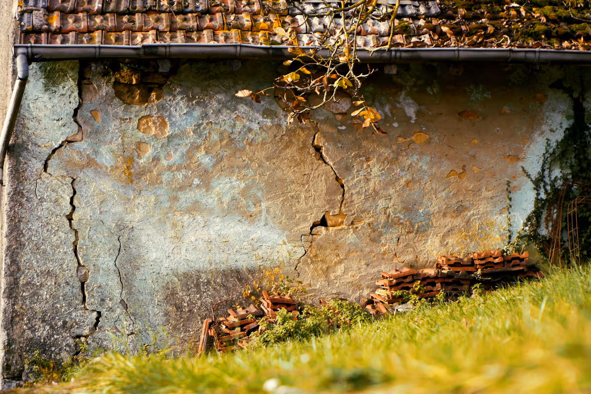 Fissure ancienne traversant l'enduit d'un mur de maison sous un rampant de tuiles, lèvres encrassées et plaques d'enduit déjà tombées autour du tracé