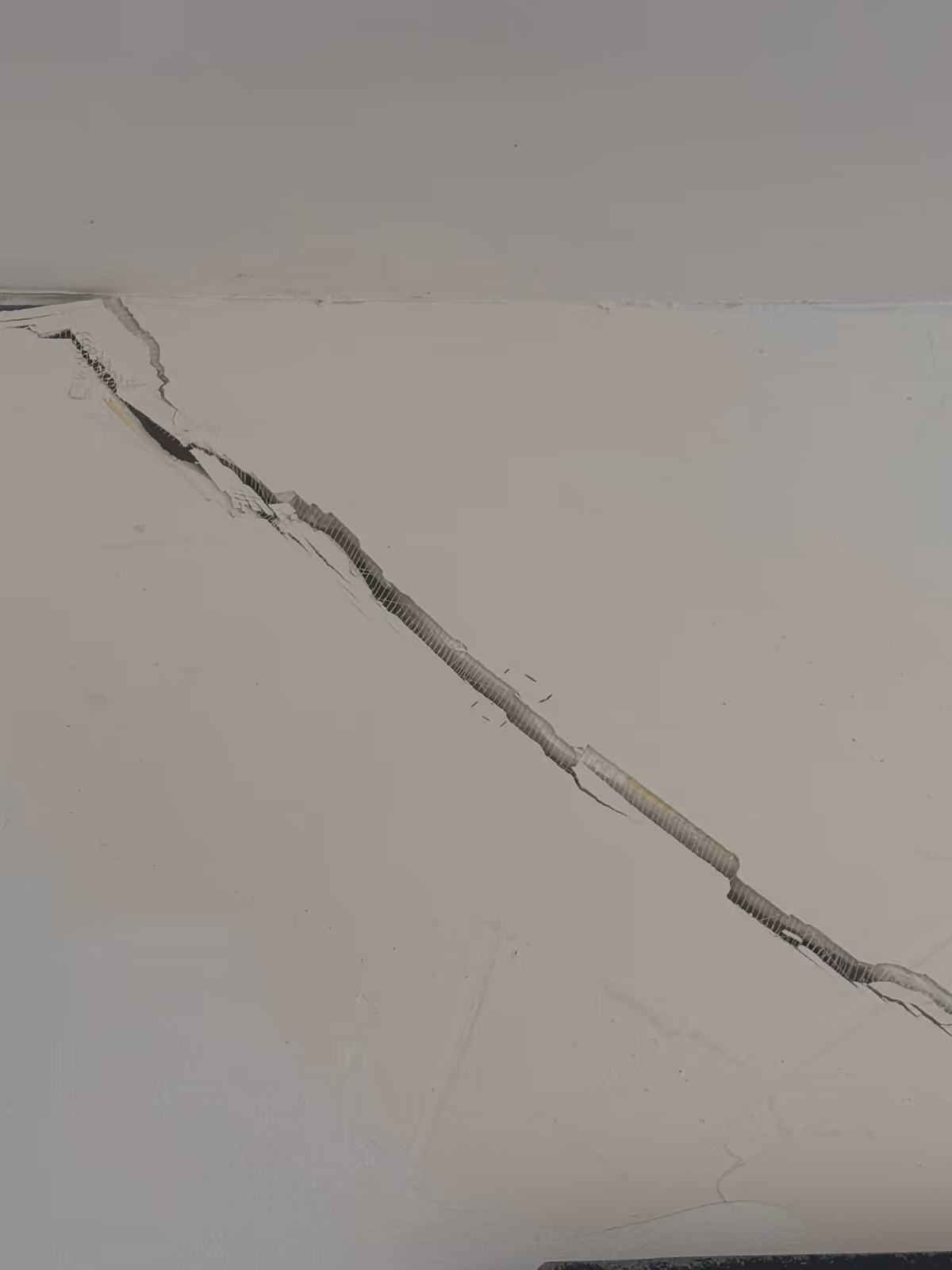 Large fissure oblique ouverte en partie haute d'un mur intérieur blanc, lèvres d'enduit décollées sur tout le tracé